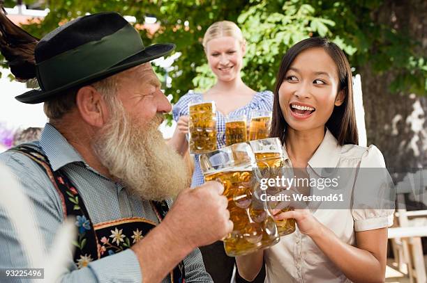 germany, bavaria, upper bavaria, bavarian man and asian woman in beer garden raising stein glasses, portrait - bayern menschen stock-fotos und bilder