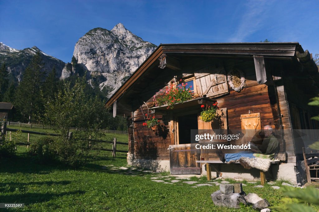 Austria, Karwendel, Senior man sitting in front of log cabin, reading a book