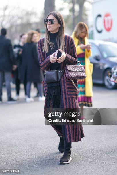 Guest wears square sunglasses, a black turtleneck, a navy blue, dark pink and yellow striped long coat with large pockets, a black studded fringe...