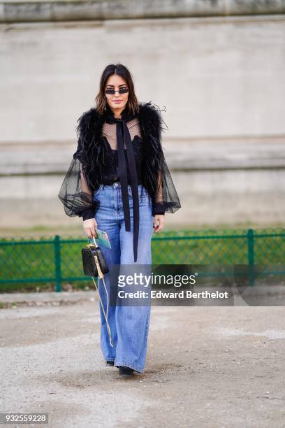 Guest wears sunglasses, a black feather boa, a black transparent tie top with long puffed sleeves, flare blue jeans, a black handbag, during Paris...