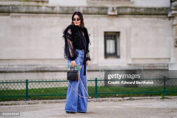 Guest wears sunglasses, a black feather boa, a black tie top with long transparent puffed sleeves, flare blue jeans, a black handbag , during Paris...