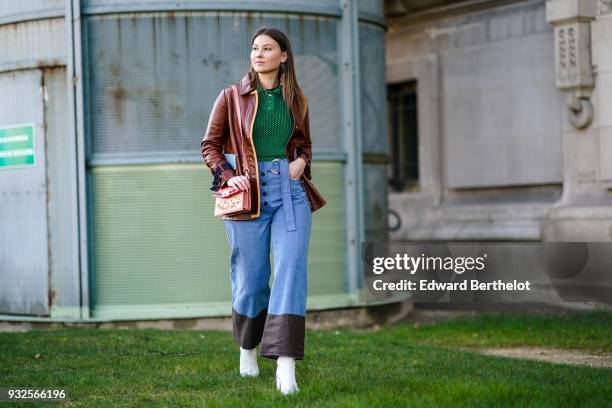 Angelica Ardasheva wears a green knit top, a camel-lined brown leather jacket, waisted blue denim button-front and sliding buckle belt wide-leg pants...