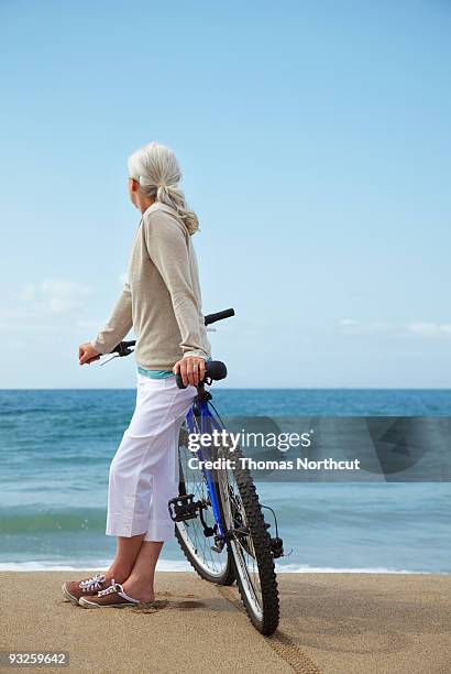 mature woman leaning against bicycle at beach. - 足首を重ねる ストックフォトと画像