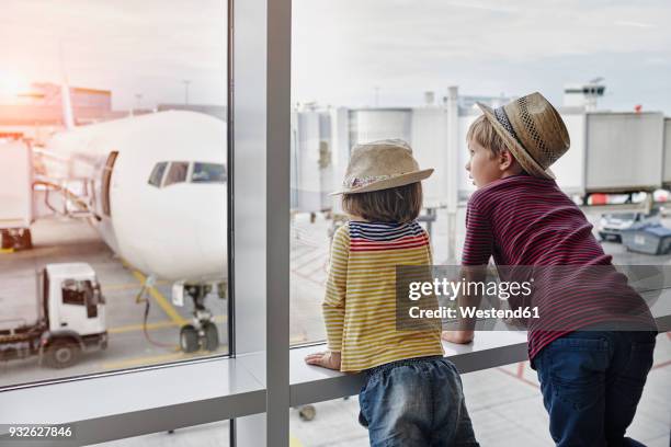 two children wearing straw hats looking through window to airplane on the apron - family travel stockfoto's en -beelden