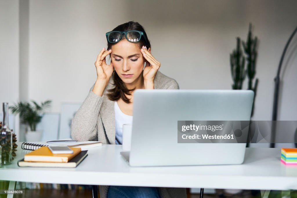 Young woman at home with laptop on desk touching her temples