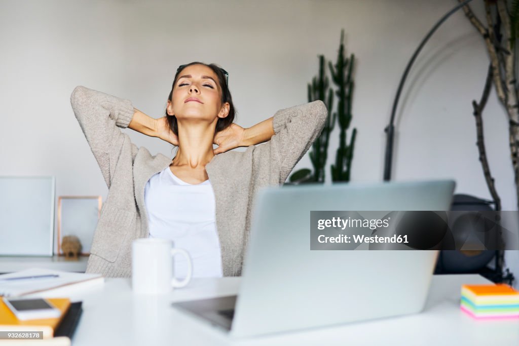 Young woman at home with laptop on desk leaning back