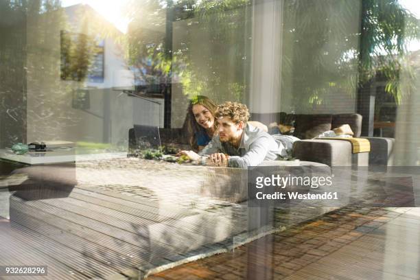 smiling couple using laptop lying on sofa at home - comfortable couple stock pictures, royalty-free photos & images