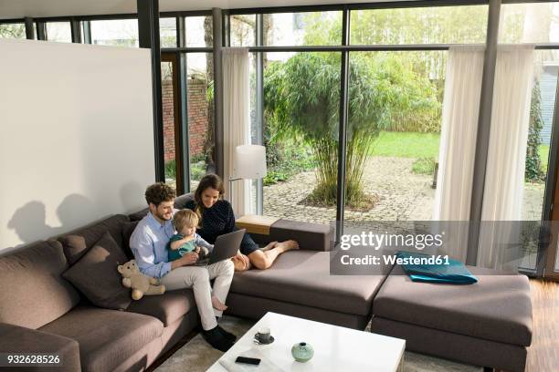 parents and son sitting on sofa in modern living room using laptop at home - familie laptop stock-fotos und bilder