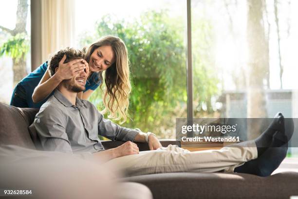 smiling woman covering her boyfriend's eyes on sofa at home - contemporary couple stock-fotos und bilder