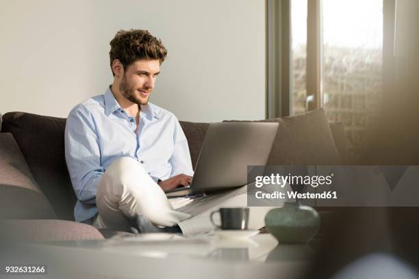 man sitting on couch at home using laptop - young man laptop couch photos et images de collection