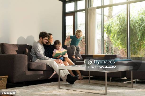 family on sofa at home reading book with boy jumping - jump dad stockfoto's en -beelden