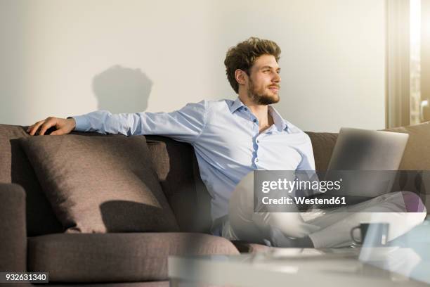 man sitting on couch at home with laptop - young man laptop couch photos et images de collection