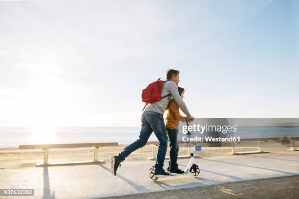 father and son riding scooter on beach promenade at sunset - holiday scooter fotografías e imágenes de stock