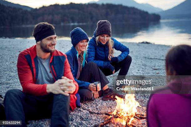 smiling friends sitting around campfire at lakeshore - bayern menschen stock-fotos und bilder