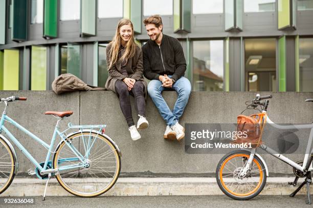 happy couple with bicycles sitting on a wall - sitzen stock-fotos und bilder