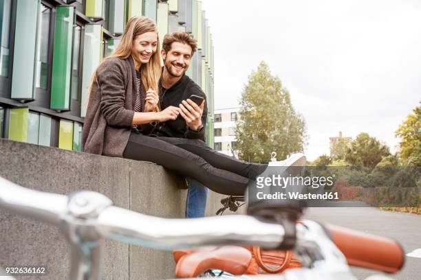 happy couple with bicycles sitting on a wall looking at cell phone - happy couple using cellphone stockfoto's en -beelden