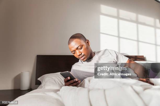 businessman waking up in bed texting on his phone in the morning. - black man sleeping in bed stock pictures, royalty-free photos & images