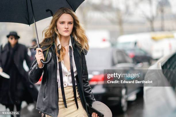 Guest holds a black umbrella and wears a white shirt with a black tie, a black leather jacket, a cream colour fringe skirt, outside Balmain, during...