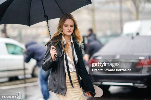 Guest holds a black umbrella and wears a black leather jacket, a white shirt with a black tie, a cream colour fringe skirt, outside Balmain, during...