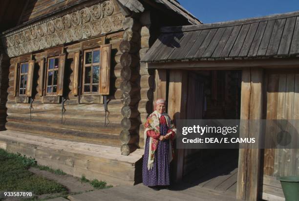 Elderly woman wearing traditional clothing in front of a peasant house, Museum of wooden architecture and peasant life, Suzdal, Russia.