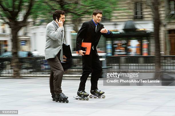 men in business attire rollerskating together along sidewalk, one phoning, the other looking at watch - roller en ligne photos et images de collection
