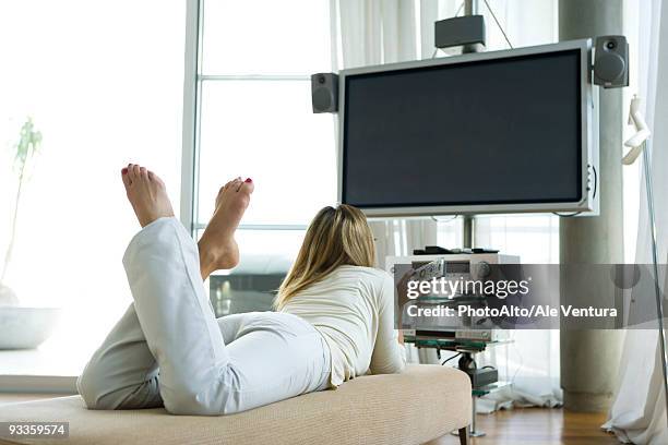 female lying on stomach watching flat screen tv with surround sound, remote control in hand, rear view - flat stomach stock-fotos und bilder