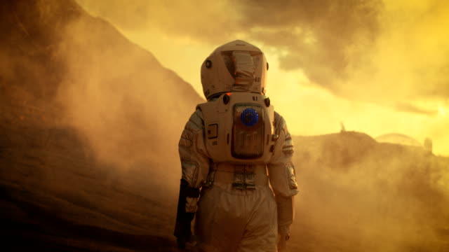 Following Shot of Female Astronaut in Space Suit Confidently Walking on Mars, Turing Around and Looking into the Camera. Red Planet Covered in Gas and Smoke.