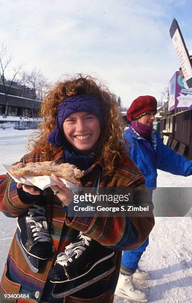 Portrait of an unidentified woman, with a pair of ice skates around her neck, who smiles broady as she holds up a beaver tail, a fried pasty, from a...