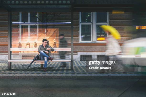 hombre en la parada de autobús en una noche de lluvia - waiting fotografías e imágenes de stock