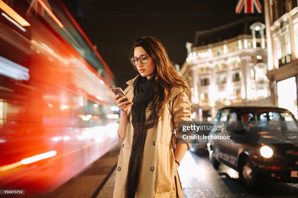 Portrait of a young woman on the busy streets of London downtown in the evening, texting for a cab