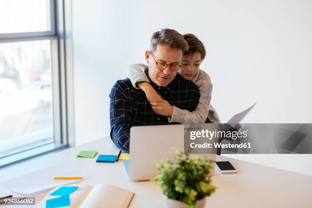 businessman using laptop at desk in office with son embracing him - dinge die zusammenpassen stock-fotos und bilder