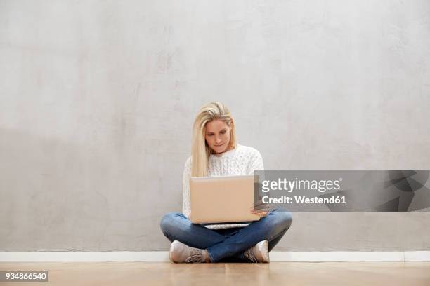 blond woman sitting on the floor in front of grey wall using laptop - schneidersitz stock-fotos und bilder