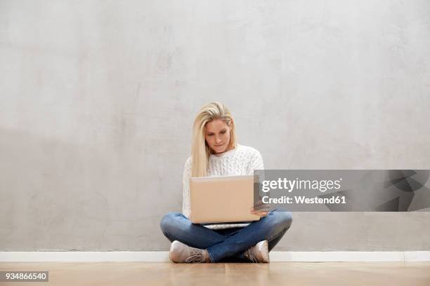 blond woman sitting on the floor in front of grey wall using laptop - gambe incrociate foto e immagini stock