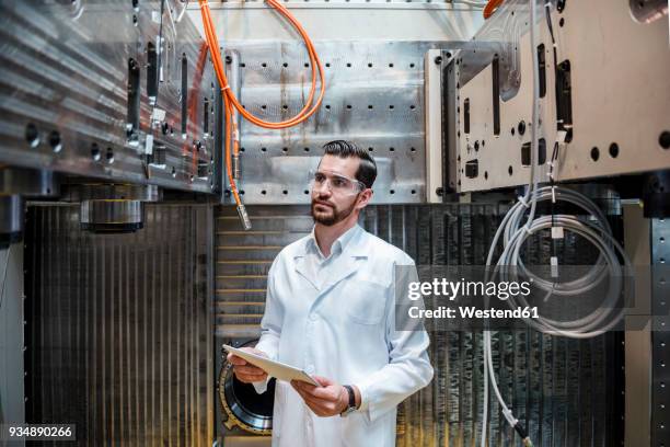 man wearing lab coat and safety goggles at machine holding tablet - scientist standing stock-fotos und bilder