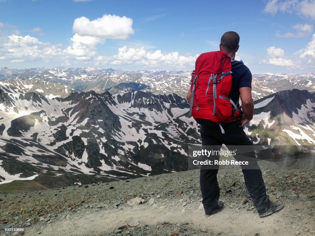 An adult male looks out over a mountain range alone on a remote mountain trail