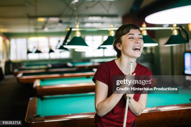 young woman laughing while holding pool cue - billar deporte de taco fotografías e imágenes de stock