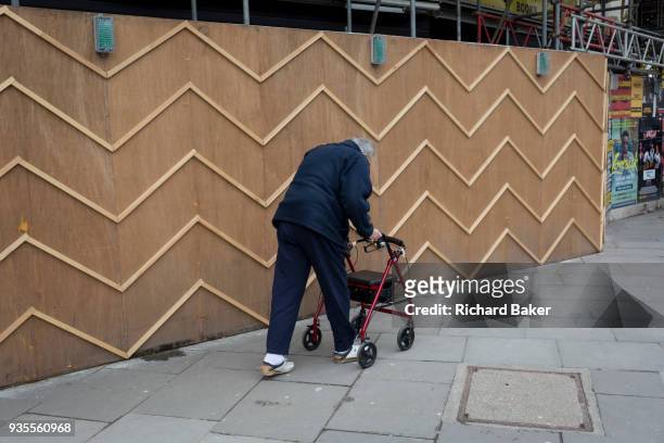 An elderly pensioner struggles to walk using a four-wheeled rollator mobility aid past the zigzag battens of a construction hoarding at Notting Hill,...