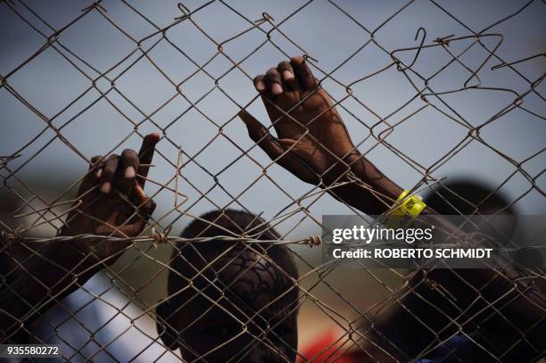 Refugee wears a bracelet that identifies him as a registered refugee looks from behind a fence as he watches others get food aid at a food...