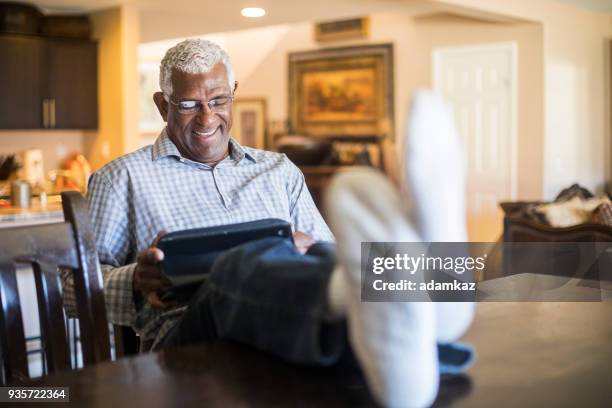 entspannte senior schwarzer mann auf tablet in küche zu hause arbeiten - black men feet stock-fotos und bilder