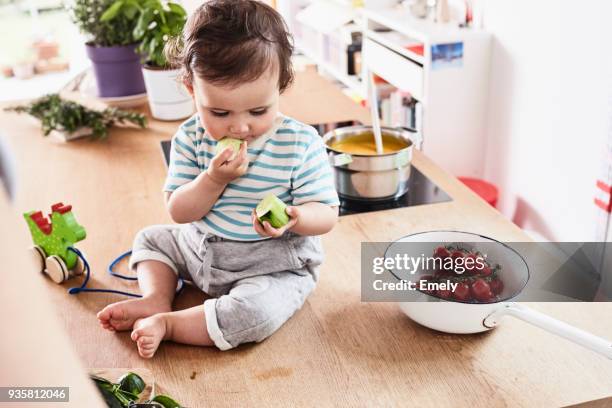 baby girl sitting on kitchen counter, eating cucumber - baby eating vegetables stock pictures, royalty-free photos & images