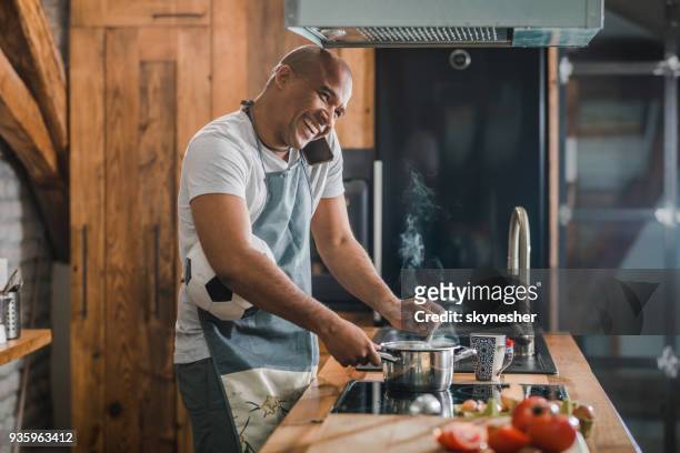 happy multi-tasking african american man talking on mobile phone while cooking in the kitchen. - multitasking man stock pictures, royalty-free photos & images