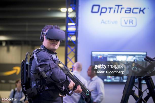 An attendee wears an Oculus VR Inc. Virtual reality headset and uses the Striker VR Arena Infinity haptic VR gun while participating in the OptiTrack...