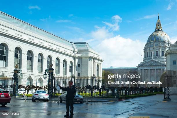 Man stands in the foreground in front of the ornate dome of San Francisco City Hall in the Civic Center neighborhood of San Francisco, California,...