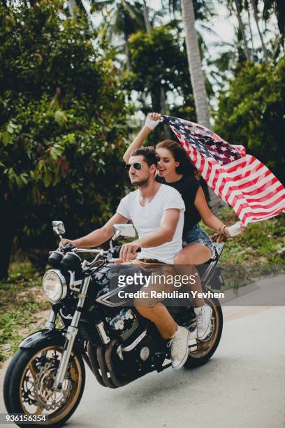 young couple holding up american flag while riding motorcycle on rural road, krabi, thailand - motorbike flag stock-fotos und bilder