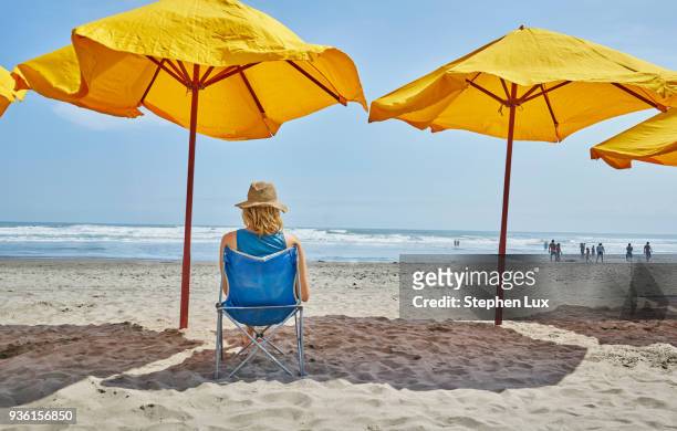 rear view of female tourist sitting under beach umbrella, camana, arequipa, peru - parasol stockfoto's en -beelden