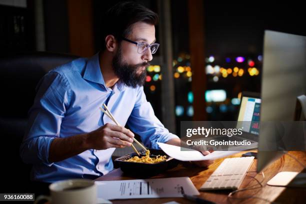 young businessman looking at computer and eating takeaway at office desk at night - working late stockfoto's en -beelden