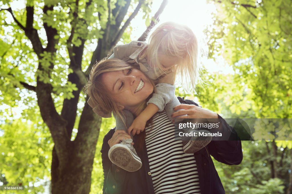 Mother giving toddler daughter shoulder carry in sunlit park