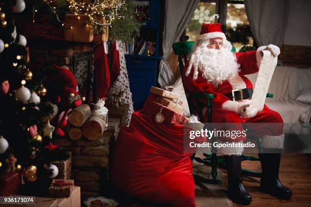 portrait of santa claus, sitting in chair with sack full of presents, looking at christmas list - papá noel fotografías e imágenes de stock
