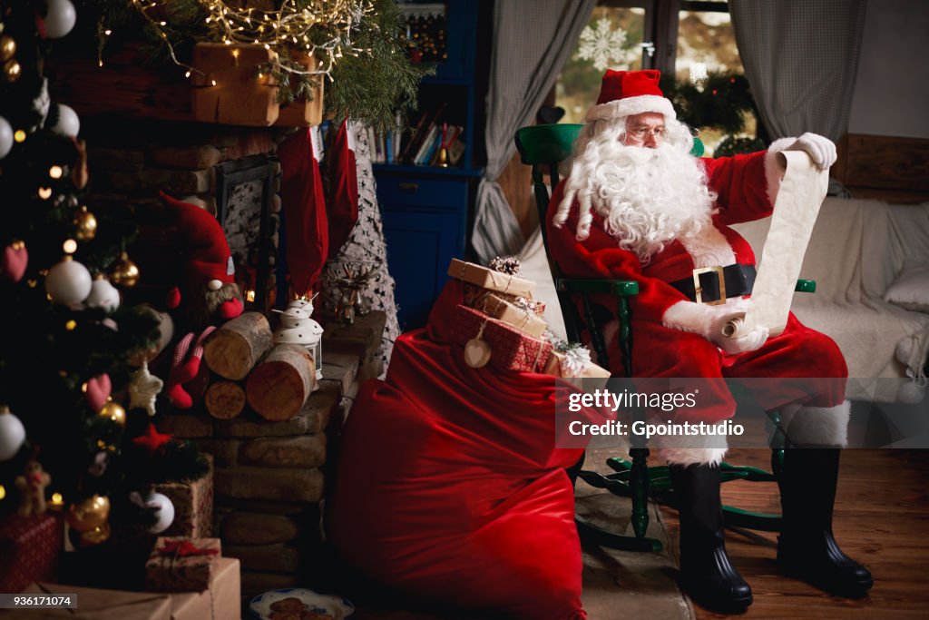 Portrait of Santa Claus, sitting in chair with sack full of presents, looking at Christmas list
