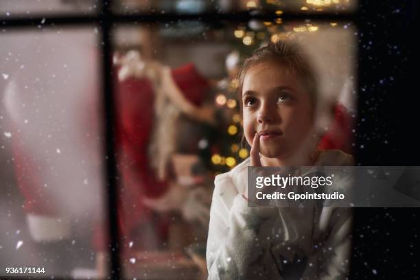 young girl looking out of window on christmas eve, santa in background leaving gifts beside tree - before christmas foto e immagini stock